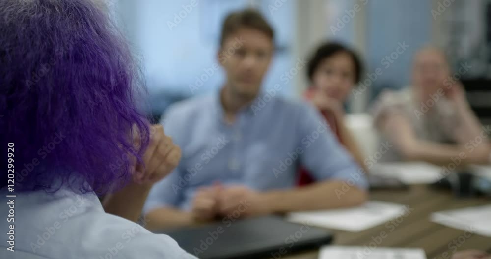 Sharp focus on foreground, looking over the shoulder of man with purple hair as he holds a meeting at wooden table with various colleagues in modern, creative office. Recorded in real time. Sharp focus on foreground, looking over the shoulder of man with purple hair as he holds a meeting at wooden table with various colleagues in modern, creative office. Recorded in real time.