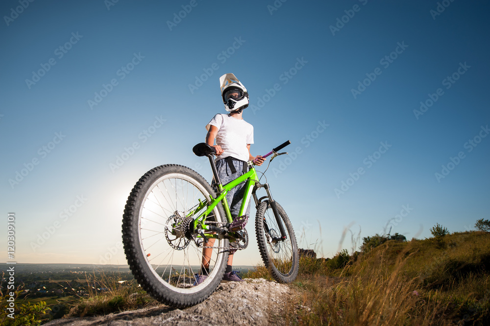 Athlete guy in helmet and glasses standing with the mountain bicycle on the hill under blue sky. Bottom view