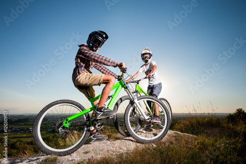 Two cyclists wearing in helmets and glasses stay on the mountain bicycles on the hill under blue sky and looking at the camera. Bottom view