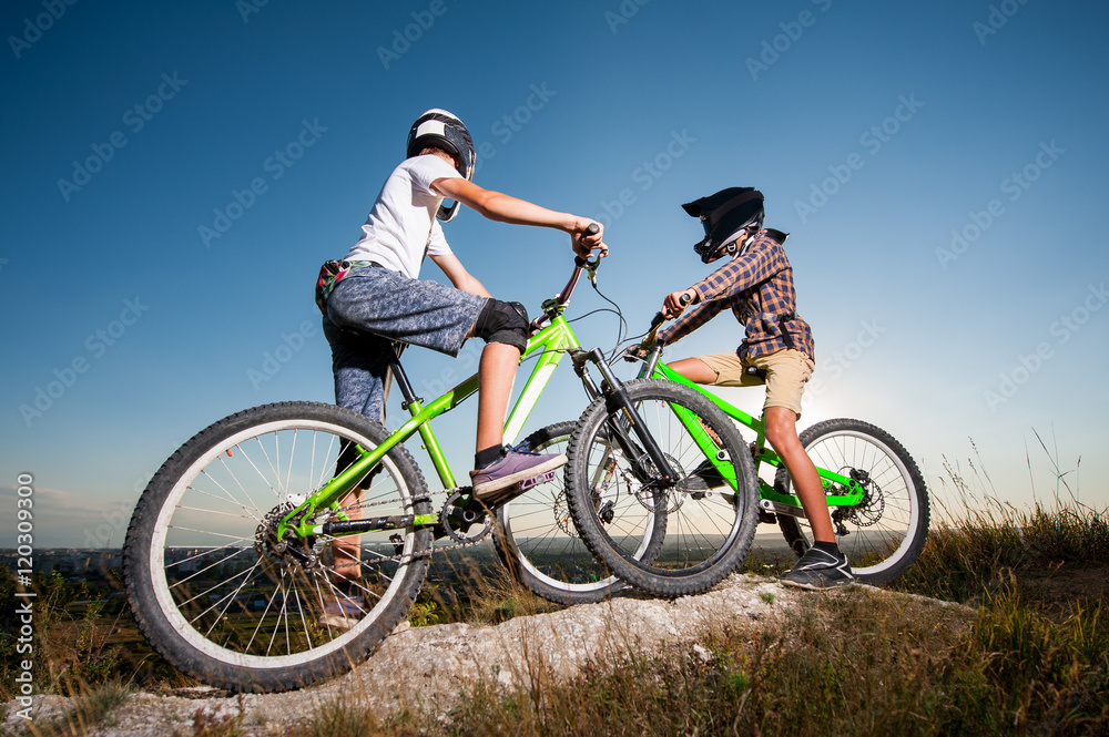 Two cyclists getting ready to riding downhill on the mountain bikes from the top of the hill under blue sky. Men looking into the distance. Bottom wide angle view Two cyclists getting ready to riding downhill on the mountain bikes from the top of the hill under blue sky. Men looking into the distance. Bottom wide angle view