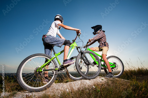 Two cyclists getting ready to riding downhill on the mountain bikes from the top of the hill under blue sky. Men looking into the distance. Bottom wide angle view