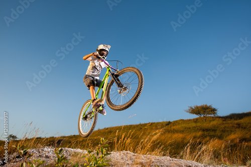 Male biker making dangerous jump on a mountain bike on the slope against blue sky. Cyclist is wearing sportswear helmet and glasses. Bottom view © anatoliy_gleb