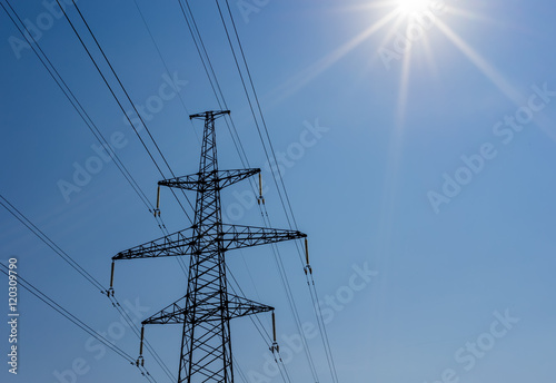 high voltage power pylons against blue sky and sun rays.
