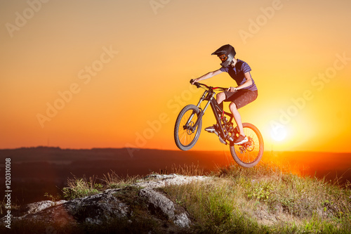 Cyclist jumping on a mountain bike on the precipice of hill against evening sky with bright sun. Cyclist is wearing sportswear helmet and glasses. Sunset