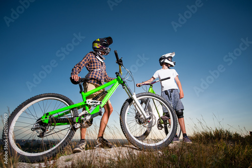 Fototapeta Naklejka Na Ścianę i Meble -  Two men wearing in helmets and glasses standing with the mountain bicycles on the hill under blue sky and looking into the distance. Wide angle view