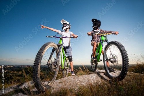 Two bicyclists wearing in helmets and glasses standing with the mountain bicycles on the hill under blue sky. Men look away and show up on something. Wide angle view © anatoliy_gleb