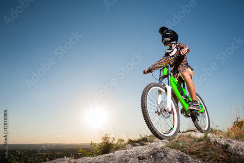 Cyclist in helmet and glasses on mountain bicycle getting ready to ride downhill from the precipice of mountain under blue sky and sun. Bottom view
