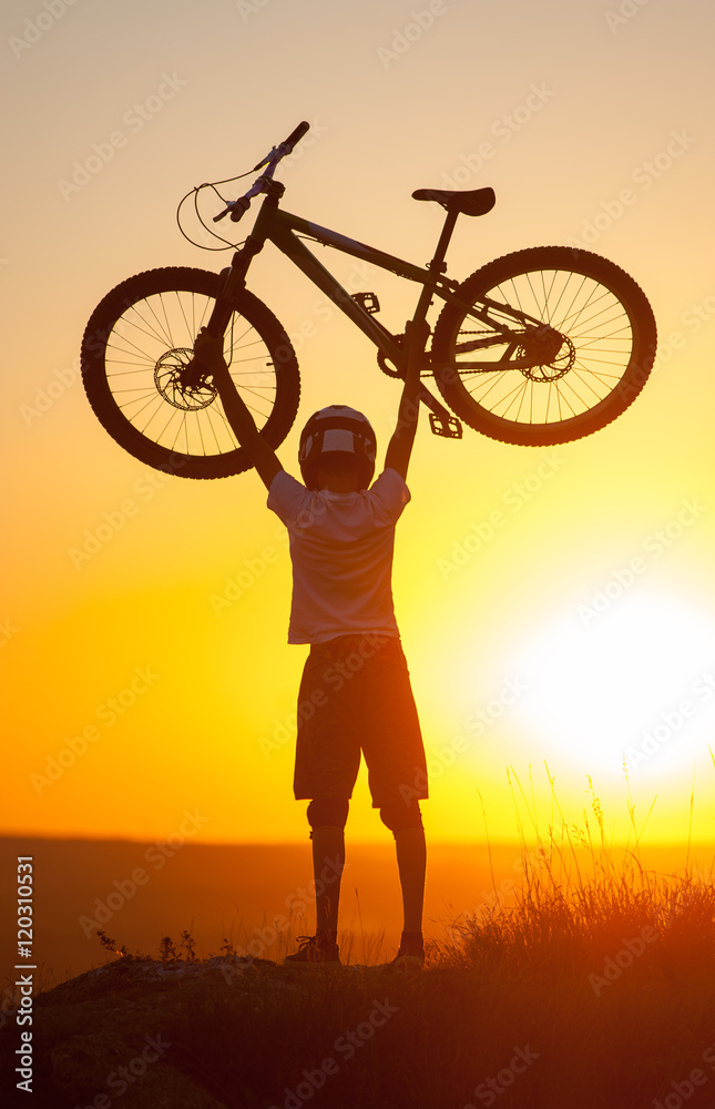 Silhouette of young cyclist in helmet holding a mountain bike over himself on the top of hill against evening sky with bright sun at the sunset Silhouette of young cyclist in helmet holding a mountain bike over himself on the top of hill against evening sky with bright sun at the sunset