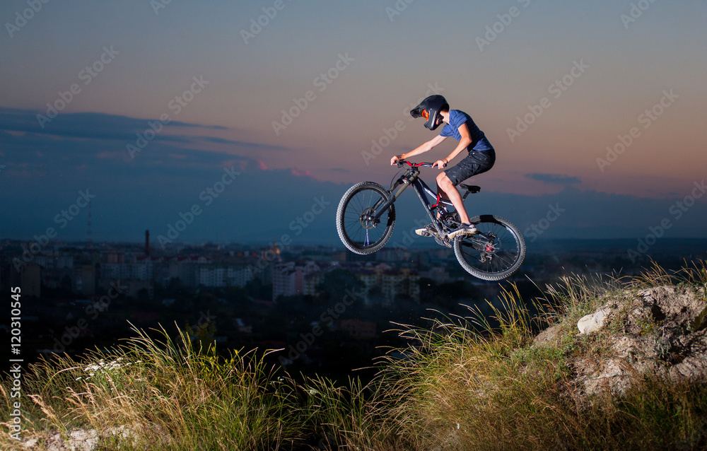 Extreme sport. Man in helmet and glasses flying on a mountain bike on the hill against evening sky and small town into the distance. Downhill cycling.