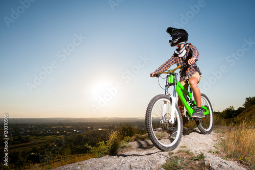 Fototapeta Naklejka Na Ścianę i Meble -  Bicyclist in helmet and glasses on mountain bike stands on the precipice of hill under blue sky against greenery and small town in the distance. Bottom view.
