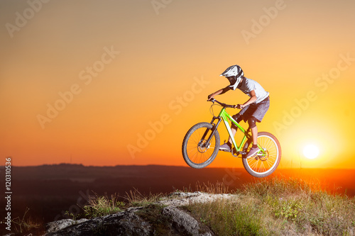 Sunset. Male biker riding downhill on a mountain bike on the precipice of hill against evening sky with bright sun. Cyclist is wearing sportswear helmet and glasses. Bottom view