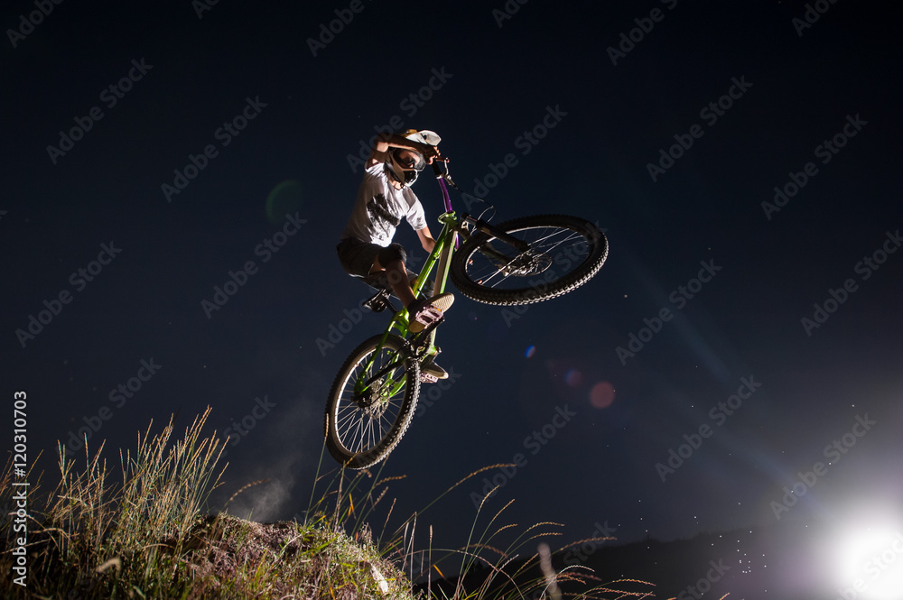 Obraz premium Bicyclist making high jump on a mountain bike on the hill against night sky. Cyclist is wearing sportswear helmet and glasses. Bottom view. Extreme downhill