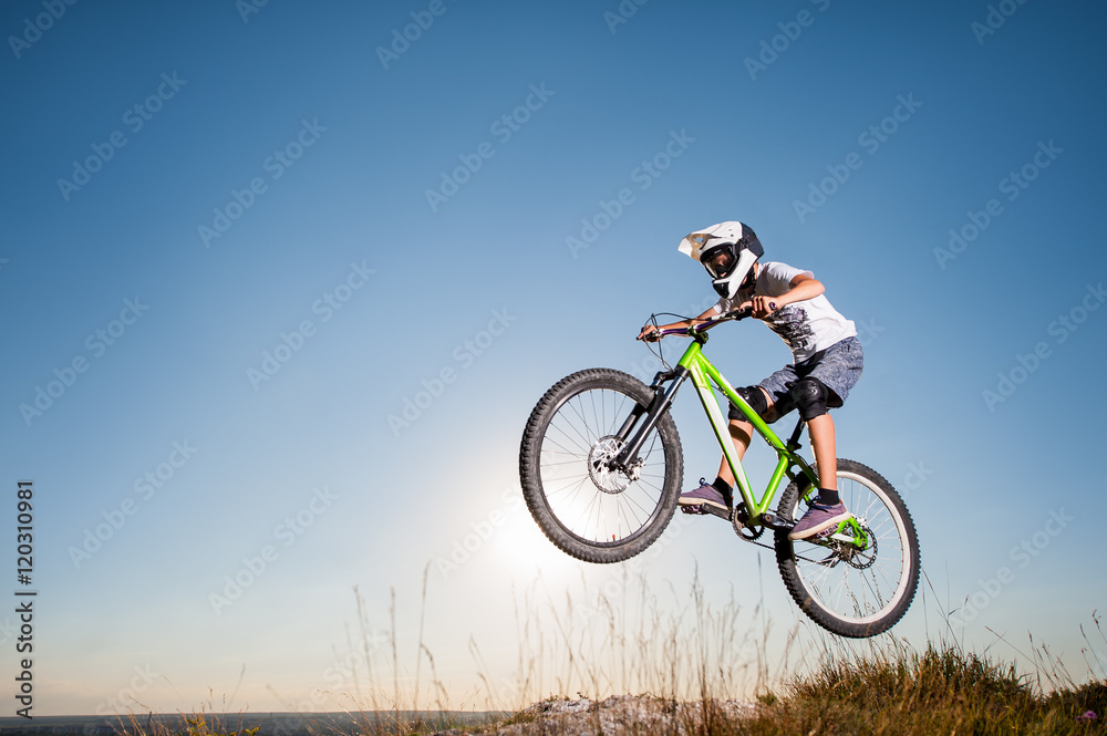 Young biker riding downhill and making dangerous jump on a mountain bike from the slope against blue sky and bright sun. Cyclist is wearing white sportswear helmet and glasses.