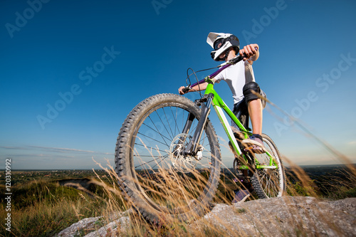 Athlete bicyclist in helmet and glasses getting ready to ride downhill on the mountain bike from the top of mountain under blue sky. Wide angle view