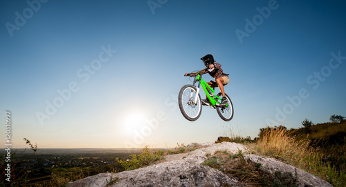 Young bicyclist in helmet and glasses making high jump on a mountain bike on the hill against blue sky. Bottom view