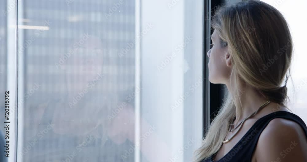 Beautiful blonde business woman looking through window and seeing her reflection in Downtown Los Angeles office with skyline visible outside. Close up, recorded hand held in real time.