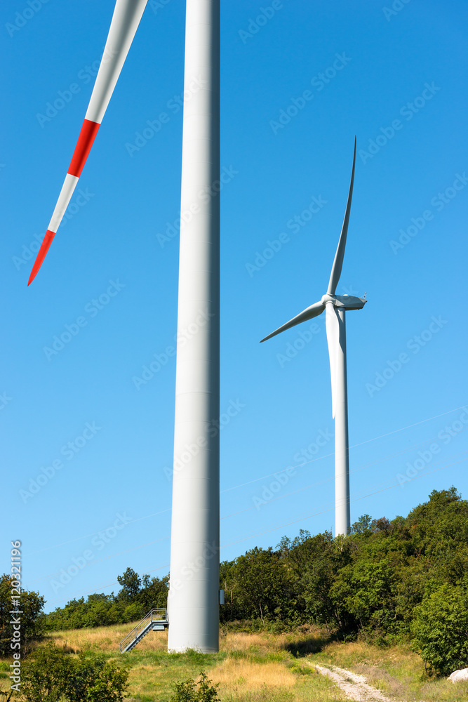 Two white and red wind turbines on a clear blue sky with a green hill Two white and red wind turbines on a clear blue sky with a green hill
