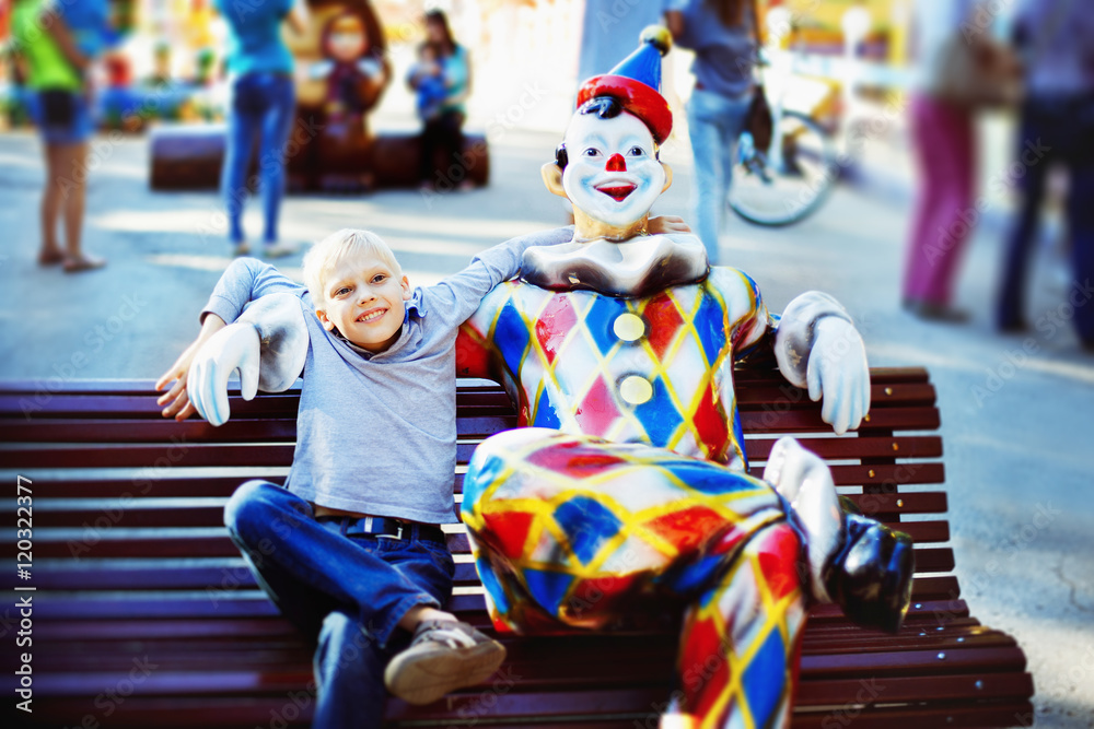 Happy boy is sitting on bench near clown figure at summer park Happy boy is sitting on bench near clown figure at summer park