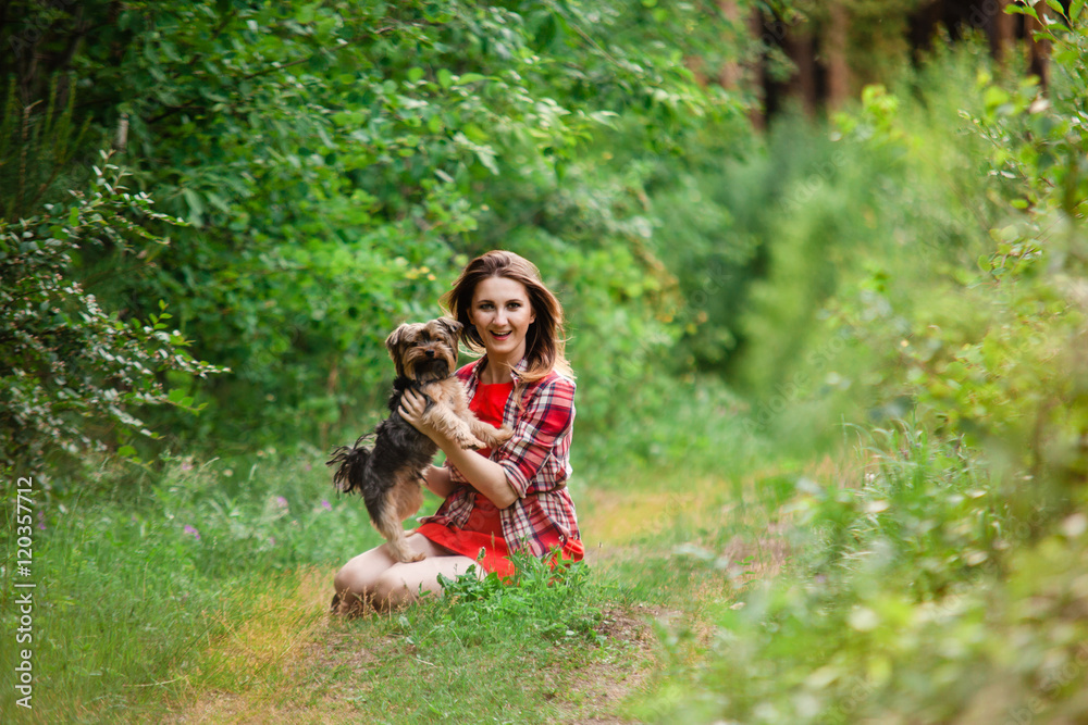 Young attractive woman in a red dress and a plaid shirt sitting in the meadow, holding her pet Yorkshire Terrier. The dog stands on its hind legs and looking at the camera. Young attractive woman in a red dress and a plaid shirt sitting in the meadow, holding her pet Yorkshire Terrier. The dog stands on its hind legs and looking at the camera.