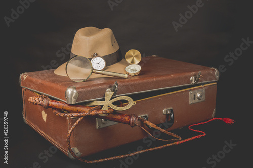 Travel and advanture concept. Vintage brown suitcase with clock, fedora hat, bullwhip, compass, magnifying glass and ankh key of life on dark background photo