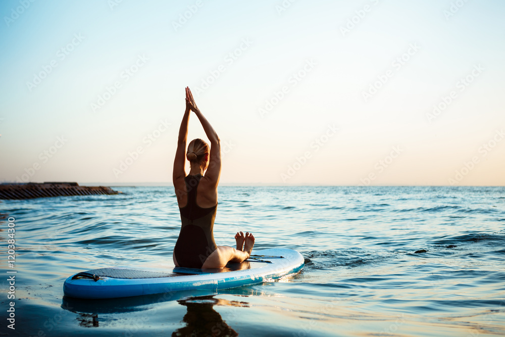 Silhouette of beautiful girl practicing yoga on surfboard at sunrise.