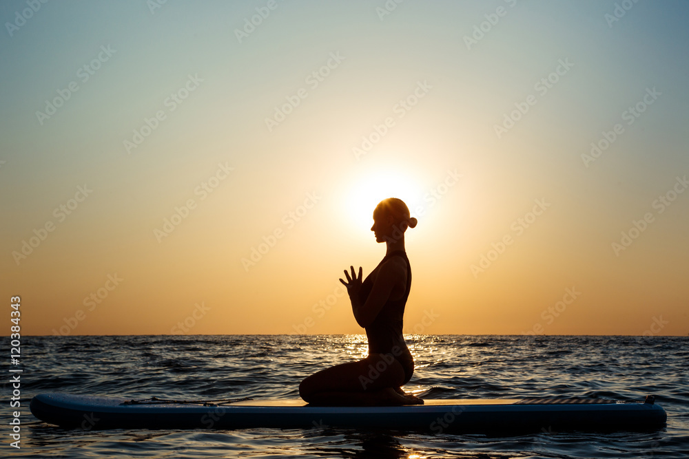 Silhouette of beautiful girl practicing yoga on surfboard at sunrise.