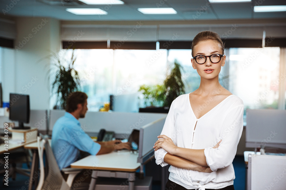 Young successful businesswoman smiling, posing with crossed arms, over office background. Young successful businesswoman smiling, posing with crossed arms, over office background.