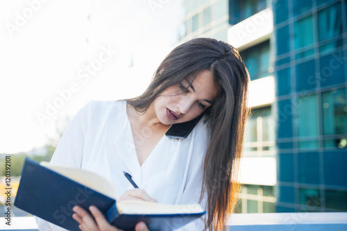 Free Download Business woman portrait outdoors talking at the phone and writing notes, with modern building as background. - GFXLoad