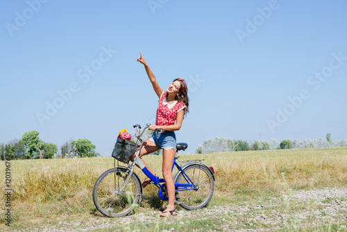 Beautiful pinup girl having fun cycling in fields under bright blue summer sky and pointing up on outdoors copy space background