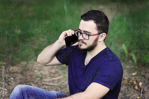 Sitting man is talking on the phone. Hi is wearing denim pants, blue t-shirt and glasses. He has got black hair, brown eyes and white skin 