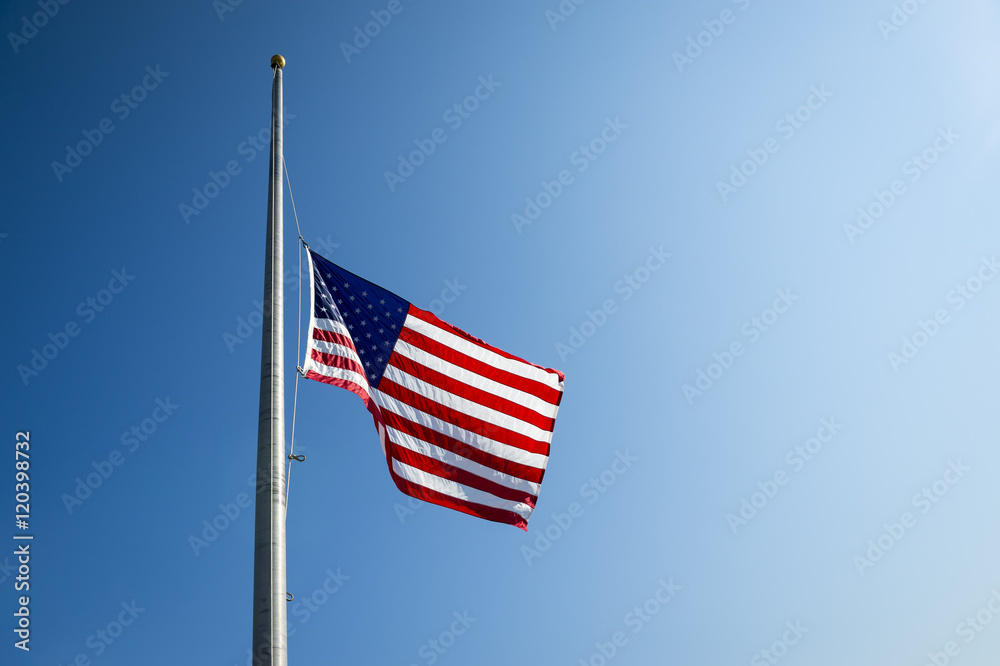 American flag flies at half mast backlit by the sun in bright blue sky