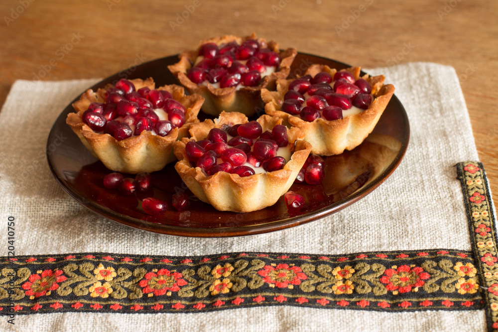 Four tartletes with custard and pomegranate in brown plate on linen tablecloth with ethnic pattern. Four tartletes with custard and pomegranate in brown plate on linen tablecloth with ethnic pattern.