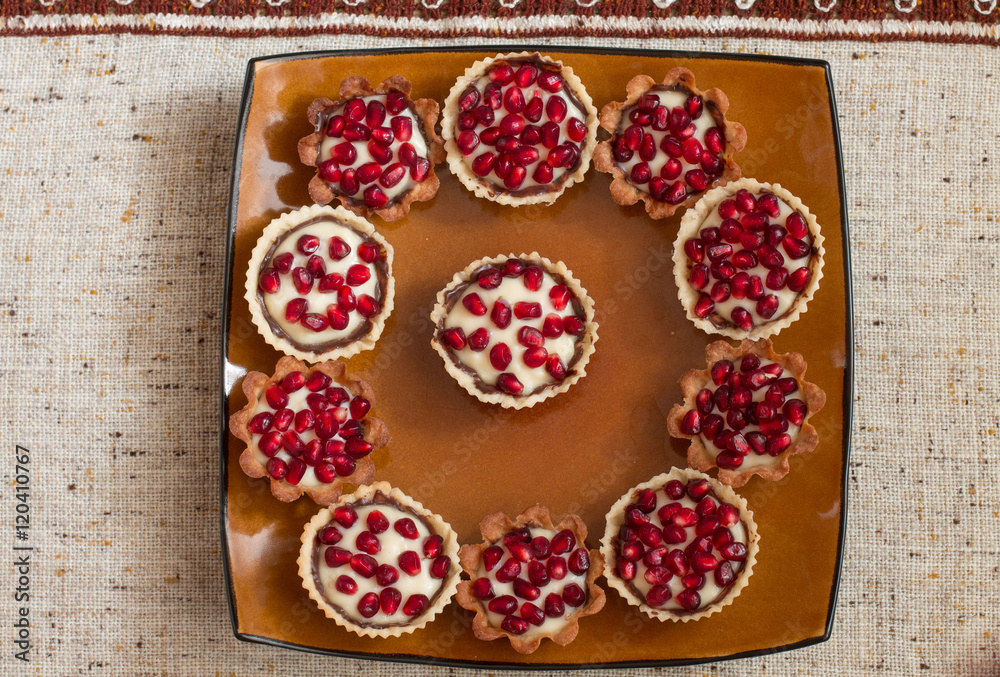 Tartletes with custard and pomegranate in big brown plate on linen tablecloth. Aerial view. Tartletes with custard and pomegranate in big brown plate on linen tablecloth. Aerial view.