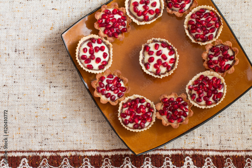 Tartletes with custard and pomegranate in big brown plate on linen tablecloth. Aerial view.