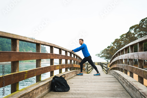 Fit sports man stretching legs before an outdoor running training on a cloudy autumn day with a sport bag on a wooden walkway. Fitness sporty man in Rodiles, Asturias.