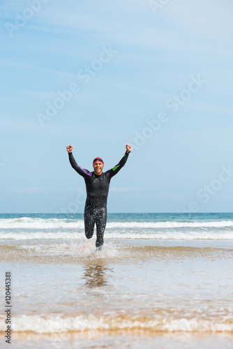 Fit sporty open water swimmer man running off shore on a beach rising arms up in victory sign after swimming triathlon competition exercise routine workout.