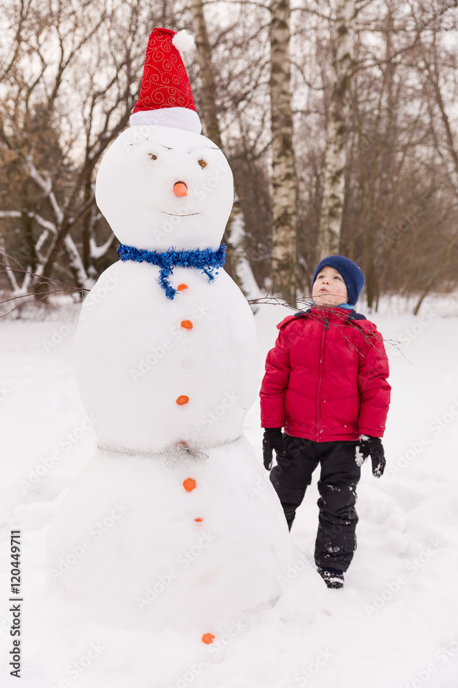 Portrait of cute kid boy with big snowman in the winter park. Child playing with snow. Lifestyle concept