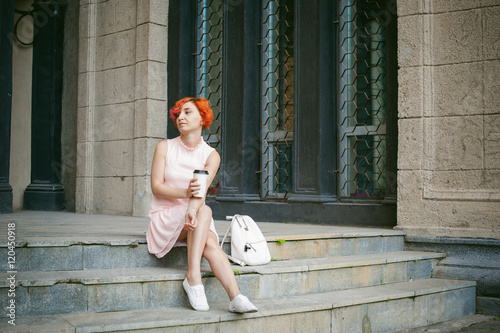 He drinks coffee from a cup. woman with dyed red hair in a pale pink dress drinking coffee, holding a paper cup of coffee to go, sitting on the steps outside a summer day.
