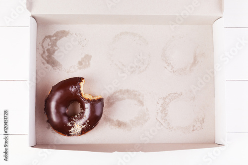 One chocolate bitten donut with sprinkles in a empty box on a white wooden background. Top view with copyspace