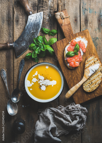 Dinner or lunch with pumpkin cream soup and salmon, ricotta and basil toasts over old rustic wooden background, top view, vertical composition