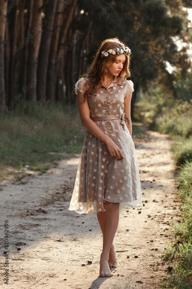 Young woman in wreath walking in forest barefoot. Beautiful girl in polka-dot dress relaxing in nature, she steps on path with pine cones, free space.