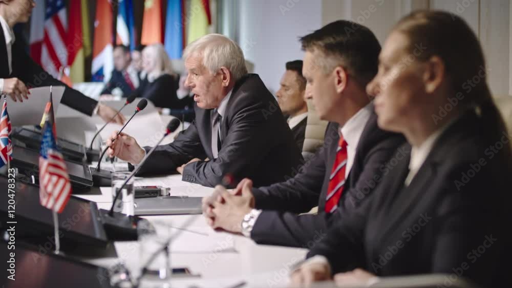 Profile shot with selective focus of senior politician sitting behind table with colleagues and talking into tabletop microphone answering question from public at press conference 