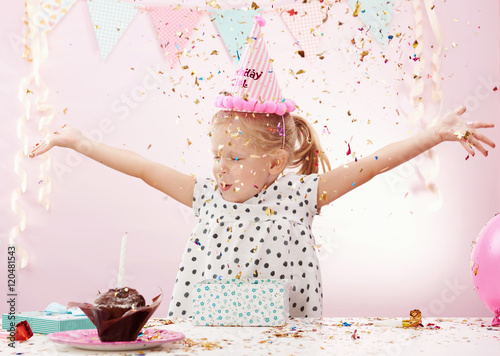 Lovely birthday girl trowing colorful confetti in the room. Cute child in pink cap having fun on party.