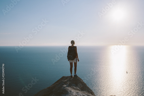 Young woman hiker with backpack standing on cliff and looking forward on the background of the sea  sky. lady tourist on top of a mountain enjoying view...