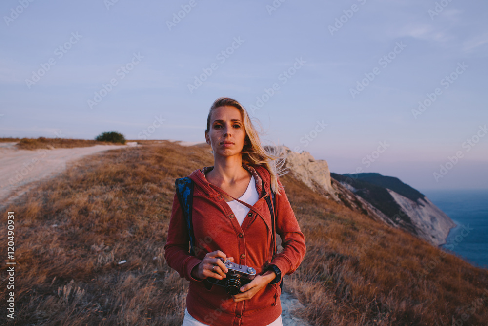 portrait of attractive young blonde woman tourist standing with an retro camera in hands on a background of mountain peaks, cliffs and the sea