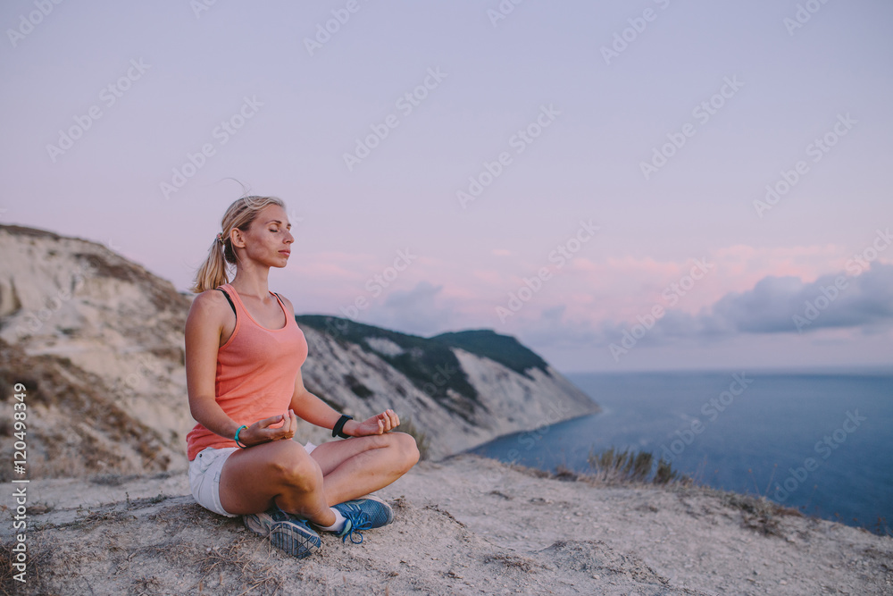 Young woman doing yoga lotus position with eyes closed outdoor on the background mountain peak cliff and sea. Sport lifestyle travel concept