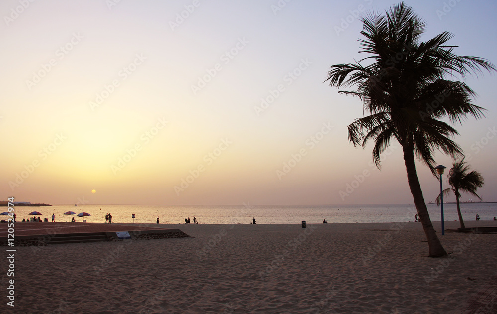 A sunset over a tropical beach with a palm tree and people silhouettes on the background