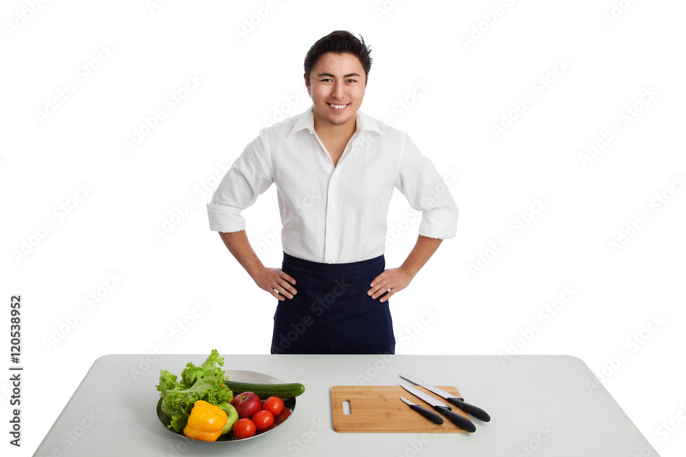 An attractive mature chef wearing a white shirt and apron, standing behind a table with kitchen knifes and mixed vegetables. White background. An attractive mature chef wearing a white shirt and apron, standing behind a table with kitchen knifes and mixed vegetables. White background.