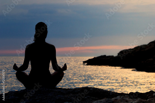 Fototapeta Naklejka Na Ścianę i Meble -  Silhouette of Woman Meditating in Lotus Position by the Sea at Sunset. Rear View. Meditation Concept.