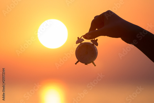 Silhouette of female hand holding small alarm clock and sunrise over sea in the background. Getting up early concept. photo